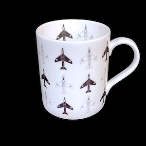 May include: White ceramic mug featuring a repeating pattern of stylized black and gray airplanes. The mug has a curved handle and a glossy finish. The design includes both detailed and outline illustrations of the aircraft.