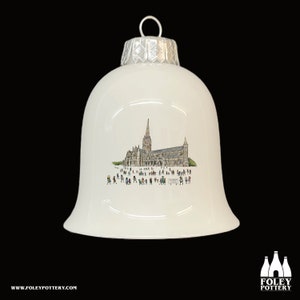 May include: White ceramic Christmas ornament shaped like a bell with a silver top. The ornament features a hand-drawn illustration of a cathedral with people walking in front of it.