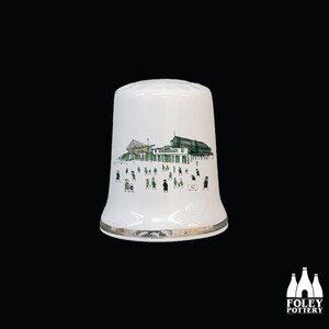 Puede incluir: Dedal de cerámica blanca con una ilustración verde y negra de un edificio y personas. La ilustración es de un estadio con gente caminando alrededor del exterior.
