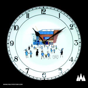 May include: A white ceramic clock with black hands and numbers. The clock face features a cartoon illustration of a football stadium with people walking towards it. The text "Highfield Road" is visible on a sign.