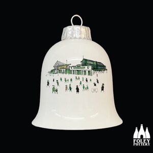 Puede incluir: Adorno navideño de cerámica blanca con forma de campana con una ilustración verde y blanca de un estadio y personas. El texto "Gate 1985" es visible en el adorno.