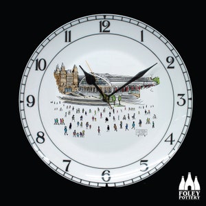 Puede incluir: Un reloj de cerámica blanca con una esfera negra y agujas negras. La esfera del reloj presenta una ilustración en blanco y negro de una estación de tren con gente caminando. La esfera del reloj también incluye el texto "LIVERPOOL LIME STREET STATION".