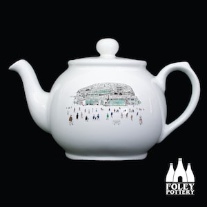 May include: White ceramic teapot featuring a black and green illustration of a building and people. The teapot has a curved spout, a rounded body, and a handle. The illustration shows a crowd of people.