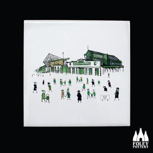 Puede incluir: Un azulejo de cerámica blanco con una ilustración en negro y verde de un estadio con gente caminando hacia él. El estadio tiene un techo verde y el texto "HOME PARK" es visible en un cartel.