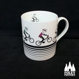 May include: White ceramic mug with a black and white striped design. The mug features a graphic of four cyclists wearing helmets and riding bicycles.