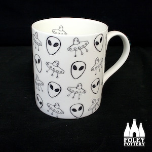 May include: White ceramic mug with a black and white pattern of aliens and flying saucers. The mug has a handle and is made by Foley Pottery.