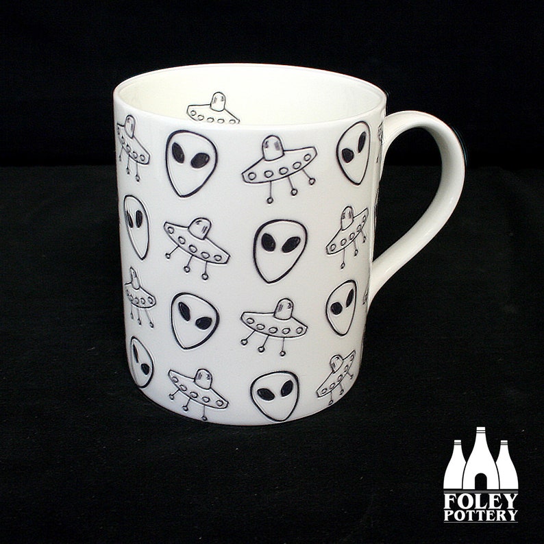 May include: White ceramic mug with a black and white pattern of aliens and flying saucers. The mug has a handle and is made by Foley Pottery.