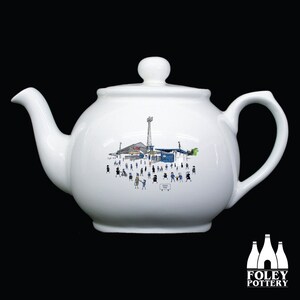 May include: White ceramic teapot featuring a black and white illustration of a stadium and people. The teapot has a round body and a spout on the left side. The handle is on the right side. The illustration depicts a stadium with a tall tower and people walking around.