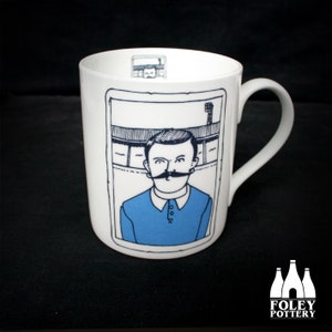 May include: White ceramic mug with a black and white illustration of a man wearing a blue jersey. The illustration is in a frame with a black border. The mug has a black handle.