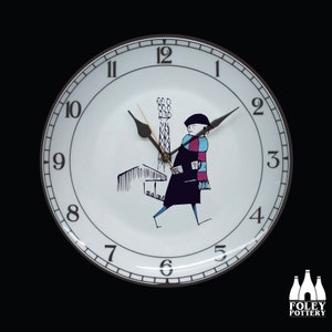 May include: A white ceramic clock face with black numbers and hands. The clock face features a black and white illustration of a person walking past a building with a tall tower. The clock face is surrounded by a brown ring with a white border.