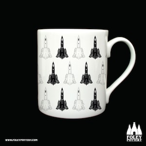 May include: White ceramic mug featuring a repeating pattern of black and white SR-71 Blackbird aircraft. The mug has a curved handle and the text "FOLEY POTTERY" and "WWW.FOLEYPOTTERY.COM" are visible.