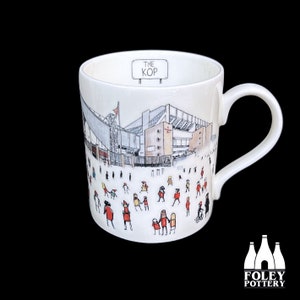 May include: White ceramic mug with a black and white illustration of a stadium and people in red and yellow. The text "THE KOP" is on a sign above the stadium.