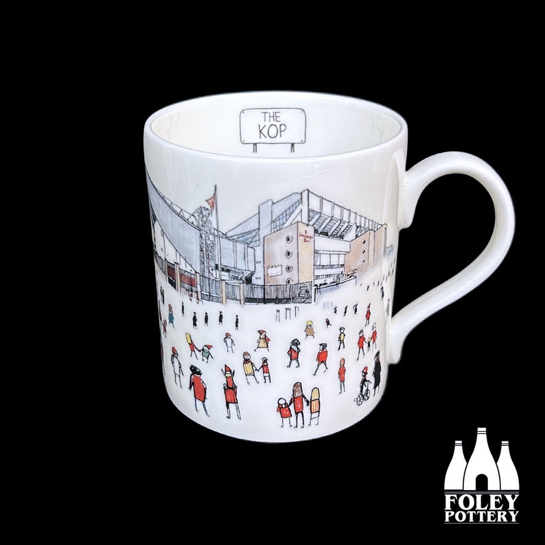 May include: White ceramic mug with a black and white illustration of a stadium and people in red and yellow. The text "THE KOP" is on a sign above the stadium.