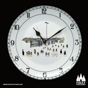 Könnte beinhalten: Eine weiße Keramikuhr mit schwarzen Zahlen und Zeigern. Das Zifferblatt der Uhr zeigt eine Schwarzweißillustration eines Gebäudes mit Personen, die davor laufen. Der Text "THE ENVIROVENT" ist auf einem Schild in der Illustration zu sehen.