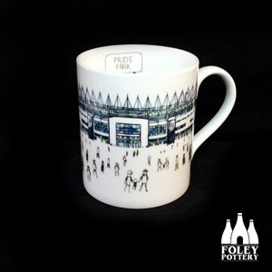 Pode incluir: Caneca de cerâmica branca com uma ilustração em preto e branco de um estádio com o texto "Pride Park" na frente. O estádio é cercado por figuras de pau de pessoas.