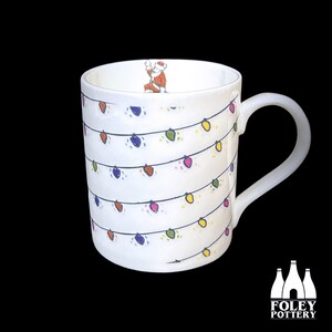 Pode incluir: Caneca de cerâmica branca com um design festivo de luzes de Natal coloridas. A caneca tem uma alça e uma pequena imagem do Papai Noel dentro.
