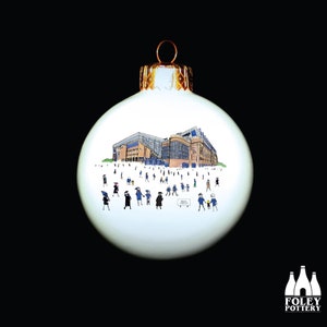 May include: White Christmas ornament with a hand-painted illustration of a stadium and people in front of it. The ornament is decorated with gold trim.