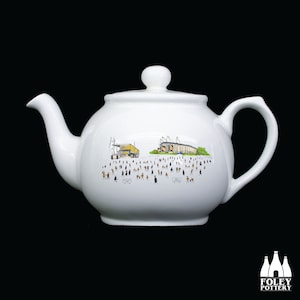 Peut inclure: Théière en céramique blanche avec une illustration de dessin animé d'un stade de football et de supporters. La théière a une poignée et un bec noirs.