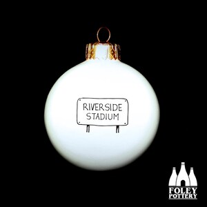 May include: White Christmas ornament with a black and white sign that reads "Riverside Stadium".