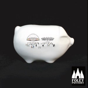 May include: White ceramic piggy bank with a black and white illustration of a stadium and people walking towards it. The piggy bank is made by Foley Pottery.