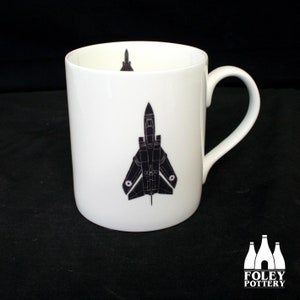 May include: White ceramic mug with a black silhouette of a fighter jet printed on the front. The jet is facing forward and has a black and white design. The mug has a black handle.
