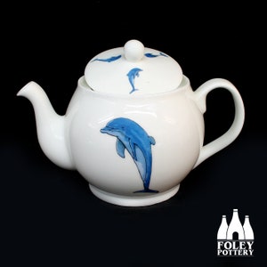 Peut inclure: Théière en porcelaine blanche avec un motif de dauphin bleu. La théière a un couvercle avec un petit bouton sur le dessus. Le dauphin saute hors de l'eau. La théière est sur un fond noir.