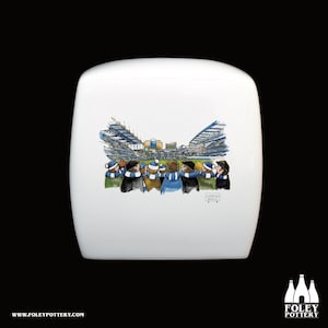 May include: White ceramic toilet tank lid with a cartoon illustration of a crowd of people watching a soccer game at Stamford Bridge. The illustration is in blue, white, and black.