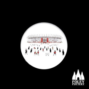 May include: A white ceramic button featuring a black and white illustration of a stadium with a red roof and people walking in front of it. The text "SUBWAY STAND" is on a sign in the foreground.