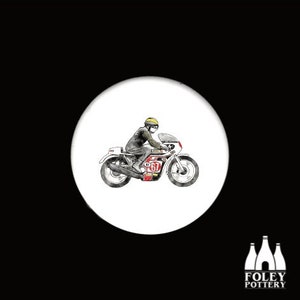 Könnte beinhalten: Ein weißer Keramik-Knopf mit einer Schwarzweiß-Illustration einer Person, die ein Motorrad fährt. Das Motorrad hat die Nummer "67" in Rot und Weiß an der Seite.