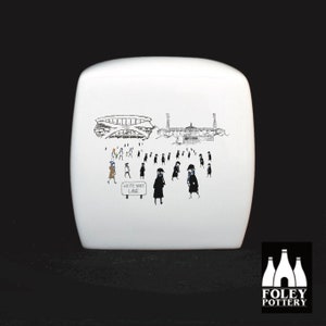 May include: White ceramic storage jar with a black and white illustration of a stadium and people walking towards it. The text "White Hart Lane" is on a sign in the image.