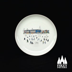 Puede incluir: Un plato de cerámica blanco con una ilustración en blanco y negro de un estadio con gente caminando alrededor. El estadio es azul y blanco con un techo rojo. El texto "Millwall FC" es visible en el plato.