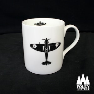 May include: White ceramic mug with a black silhouette of a Spitfire airplane on the front. The airplane is facing forward and has a black and white design. The mug has a black handle.