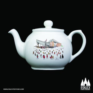 May include: White ceramic teapot featuring a cartoon illustration of a stadium and people. The stadium is a red brick building with a grey metal roof. The people are all wearing red and white clothing. The text "The Kop" is visible on the teapot.