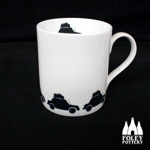 May include: White ceramic mug featuring a black silhouette of a car with luggage on the roof. The mug has a black handle and the design is repeated around the mug.
