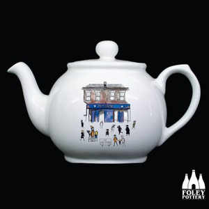 May include: White ceramic teapot with a whimsical illustration of a building with the text "OAK STAND" and figures of people. The teapot has a spout, handle, and lid. The Foley Pottery logo is at the bottom right.