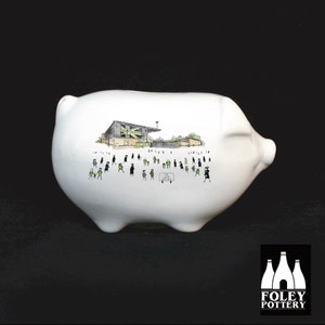 May include: White ceramic piggy bank with a black and white illustration of a soccer stadium and people in the stands. The text "The Ground" is on a sign in the illustration. The piggy bank is made by Foley Pottery.