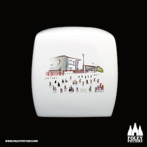 Puede incluir: Taza de cerámica blanca con una colorida ilustración de un estadio y una multitud de personas. El estadio tiene detalles en rojo y blanco. El texto "KINGSHOLM STADIUM" es visible. La taza tiene el logotipo y la dirección del sitio web de Foley Pottery.
