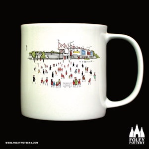 Könnte beinhalten: Weiße Keramiktasse mit einer Schwarzweißillustration eines Stadions mit Menschen in rot und blauer Kleidung. Der Text "Broadfield Stadium" ist auf die Tasse gedruckt.