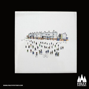 May include: A white ceramic tile featuring a hand-drawn illustration of a stadium with a crowd of people in front of it. The stadium is blue and white with a brown roof. The people are all wearing different coloured clothes. The tile has the words "Hillsborough Jones" written on a sign in the foreground.