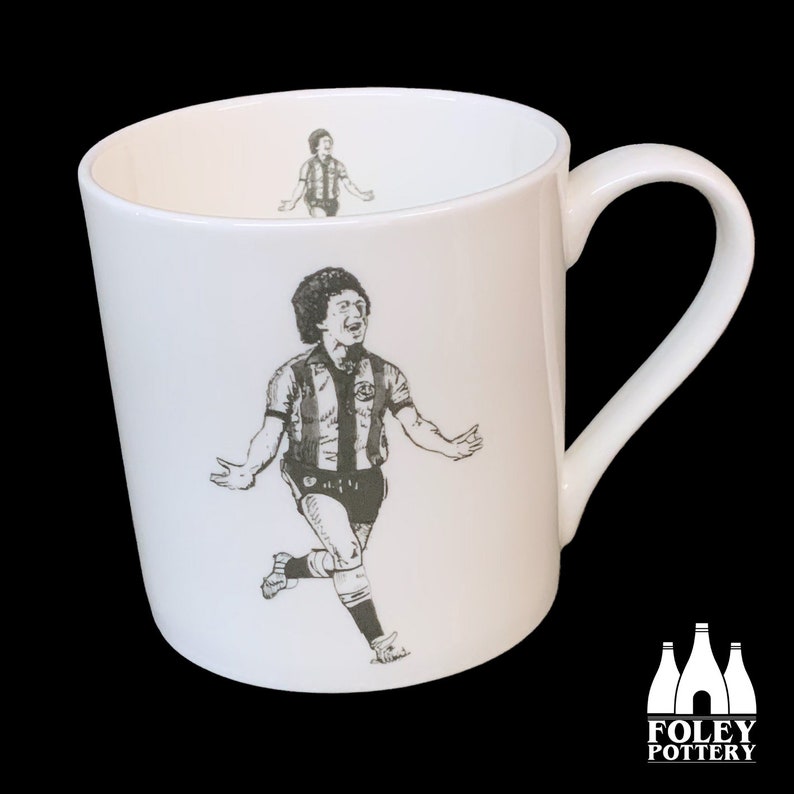 May include: White ceramic mug featuring a black and white illustration of a football player running and celebrating. The mug has a handle and the words "FOLEY POTTERY" are printed on the bottom.