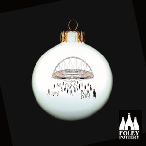 May include: White Christmas ornament with a black and white illustration of Wembley Stadium and people walking towards it. The ornament has a gold top and the text "Wembley Stadium" is visible on a sign.