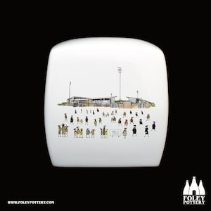 Könnte beinhalten: Weiße Keramikvase mit einer handgezeichneten Illustration einer Stadionszene, die eine Menschenmenge und den Text "PRELLI STADIUM" zeigt. Die Vase hat eine abgerundete rechteckige Form und das Foley Pottery-Logo.