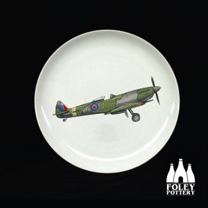 Op de afbeelding: Een wit keramiek bord met een groene en grijze illustratie van een Brits Supermarine Spitfire-vliegtuig uit de Tweede Wereldoorlog. Het vliegtuig heeft de tekst "RW385" en "U4" aan de zijkant. Het bord is gemaakt door Foley Pottery.