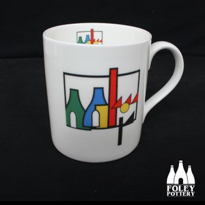 May include: White ceramic mug with a geometric design in red, yellow, blue, green, and black. The design features a stylized cityscape with buildings and a sun. The mug has a black handle and the words "FOLEY POTTERY" are printed on the bottom.