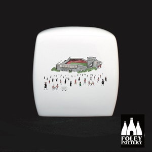 May include: White ceramic box with a black and white illustration of a stadium with people in the foreground. The stadium has a red field and a gray roof. The text "FOLEY POTTERY" is printed in black on the bottom of the box.