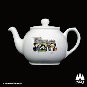 May include: White ceramic teapot with a black and white cartoon illustration of a crowd of people watching a soccer game. The teapot has a lid and a handle.