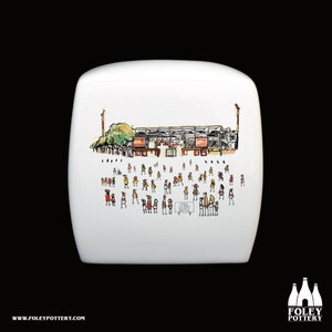May include: White ceramic piggy bank with a hand-painted illustration of a stadium with people in the stands. The stadium is red and yellow with a green lawn. The text "Stade de la Butte" is visible on the bank.