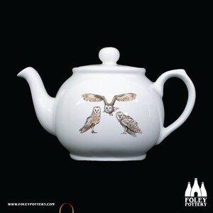 Puede incluir: Tetera de cerámica blanca con un diseño de tres búhos comunes. Los búhos están en varias poses, incluyendo uno volando con las alas extendidas. La tetera tiene un cuerpo redondo y un pico en el lado izquierdo.