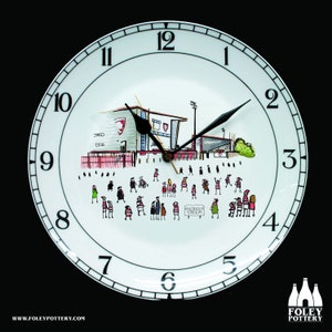 Puede incluir: Un reloj de cerámica blanca con números y manecillas negros. La esfera del reloj presenta una ilustración de dibujos animados de un estadio con gente en las gradas. El texto "KINGSHOLM STADIUM" está impreso en la esfera del reloj.