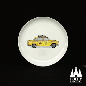 Può includere: Un piatto in ceramica bianca con un'illustrazione di un taxi giallo. Il taxi ha un tetto a scacchi bianco e nero e le parole "NYC TAXI" sul lato.